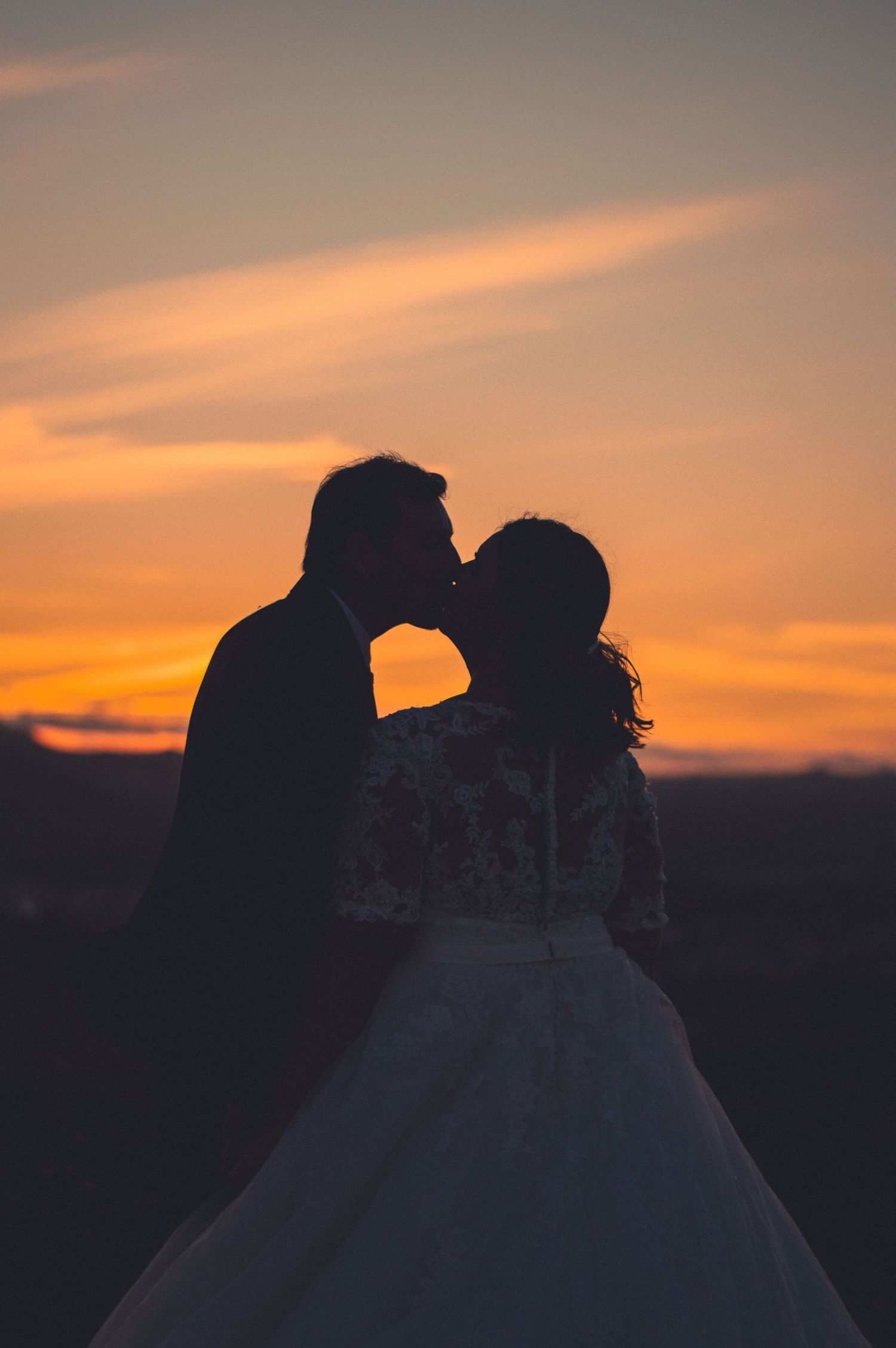 Silhouette of newlyweds kissing against dramatic orange sunset sky.