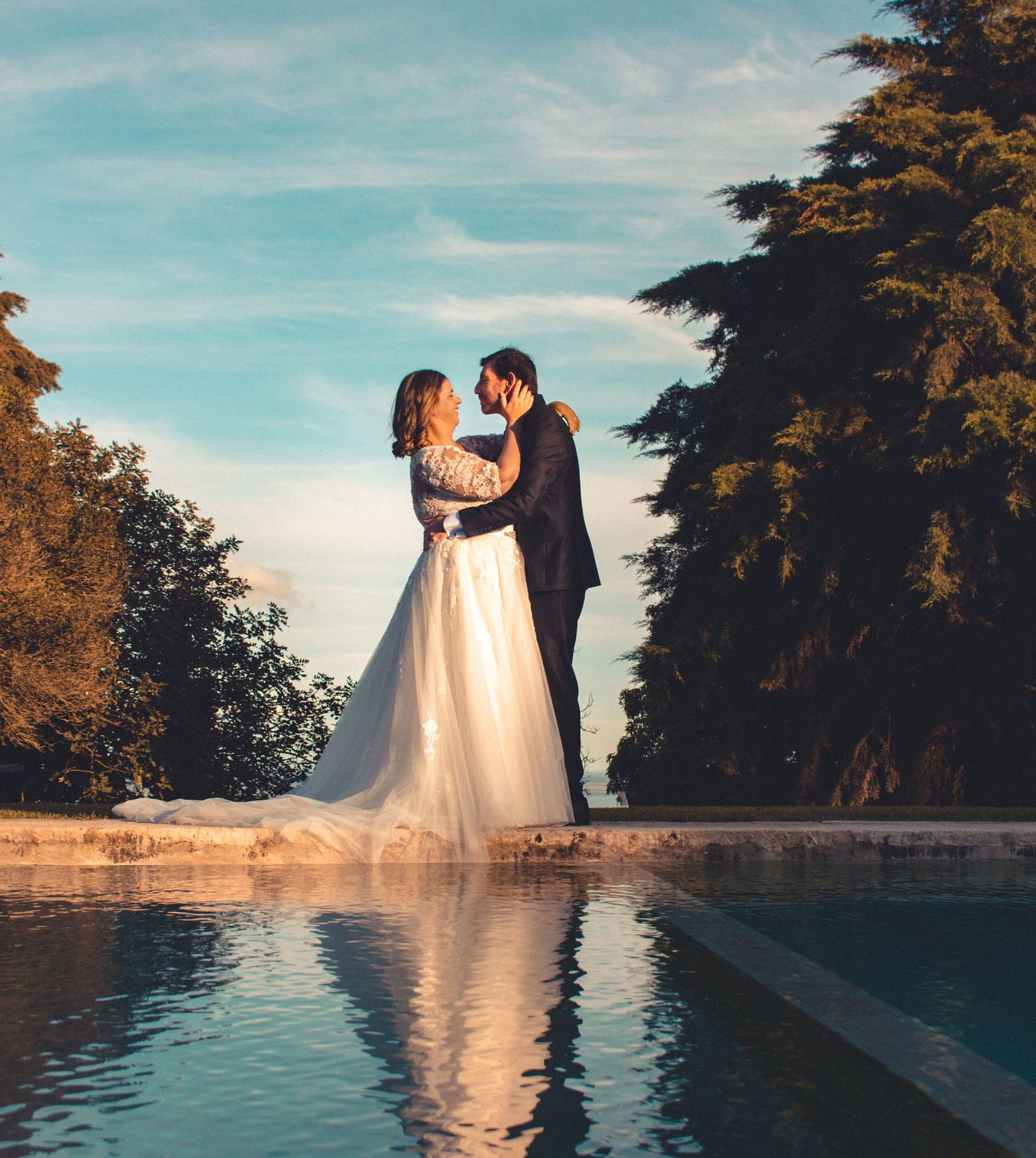 Wedding couple embracing by a reflective pool during golden hour with trees in the background.