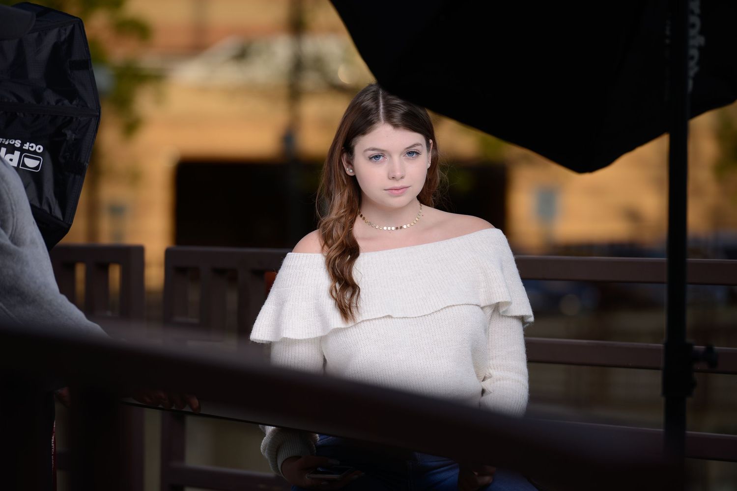 Portrait in an off-shoulder white sweater against a blurred outdoor background with railing in evening light.