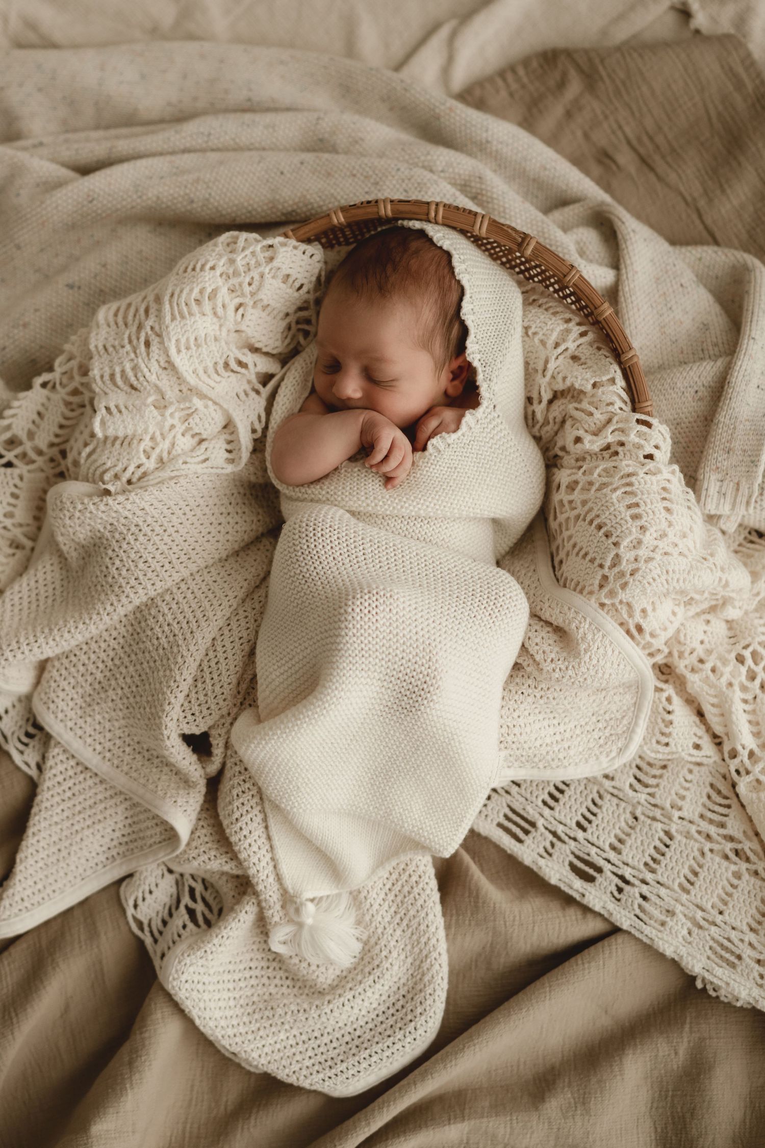 Sleeping newborn wrapped in white knit blanket nestled in textured cream fabrics.