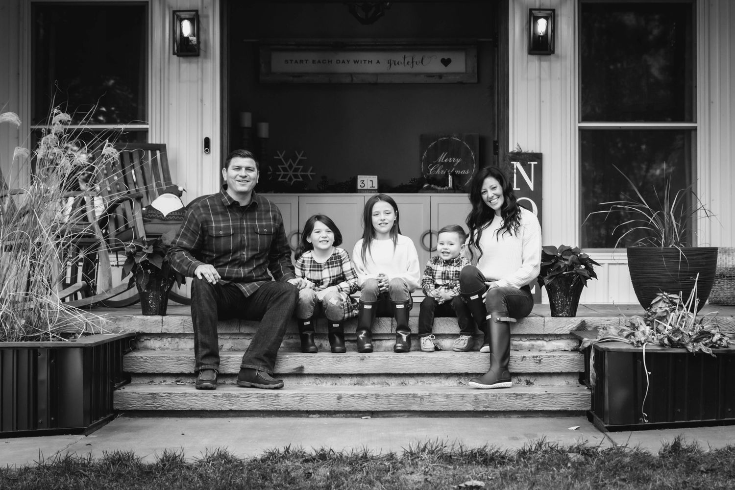 A family sitting together on the front porch steps of a house with potted plants on either side.
