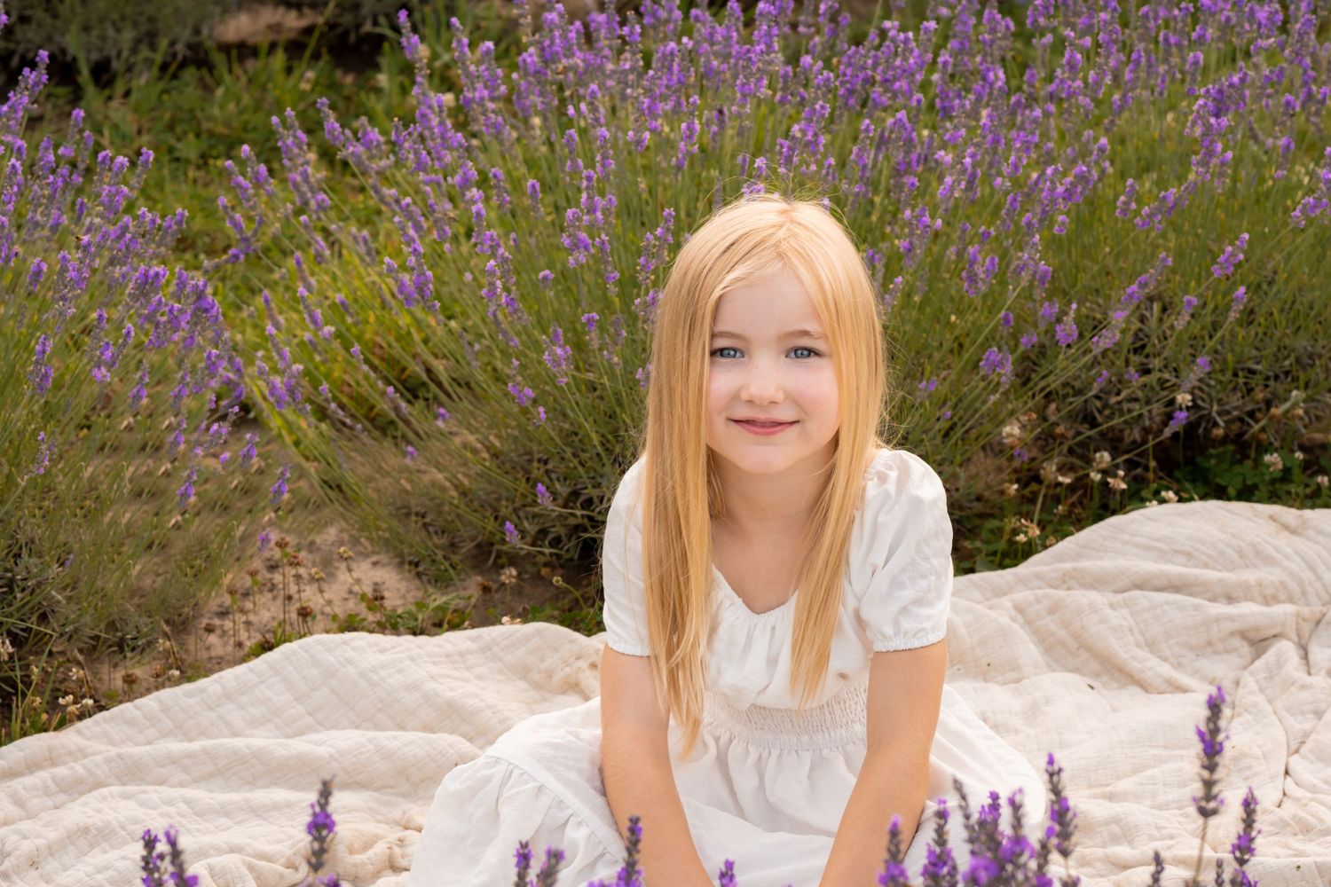 Young girl in white dress sitting among blooming lavender flowers.