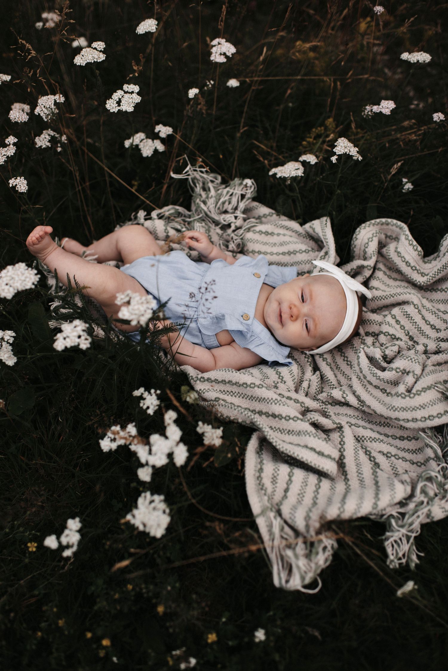 Infant in blue outfit rests on striped blanket amid white flowers in artistic outdoor portrait.