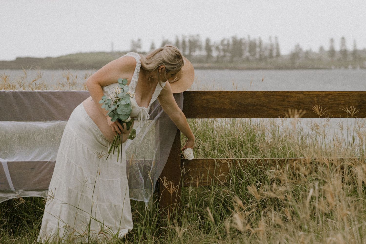 A bride in a white dress leans against a rustic wooden fence surrounded by tall grass at sunset.