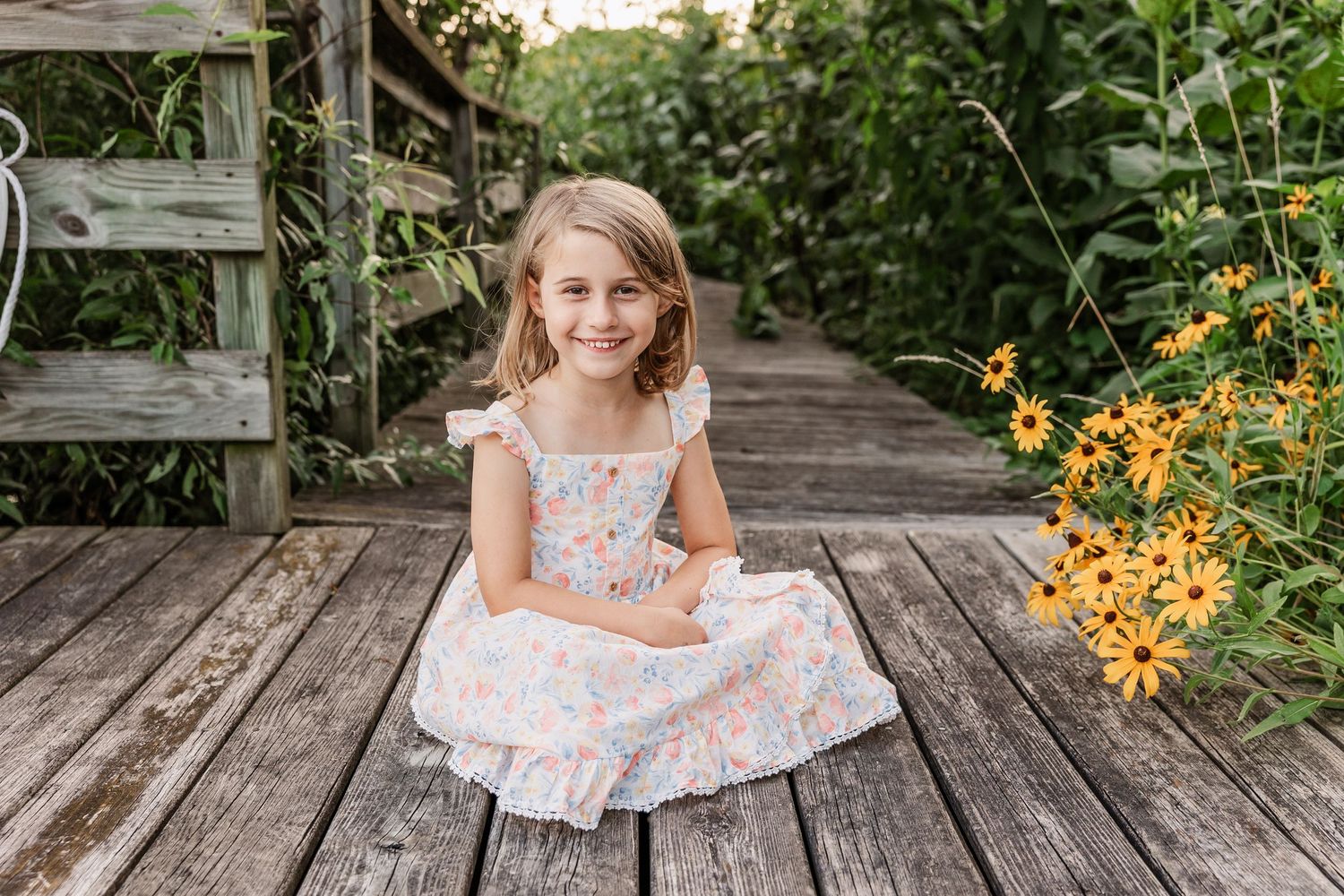 Young person in floral dress sitting on wooden boardwalk with yellow flowers in background.