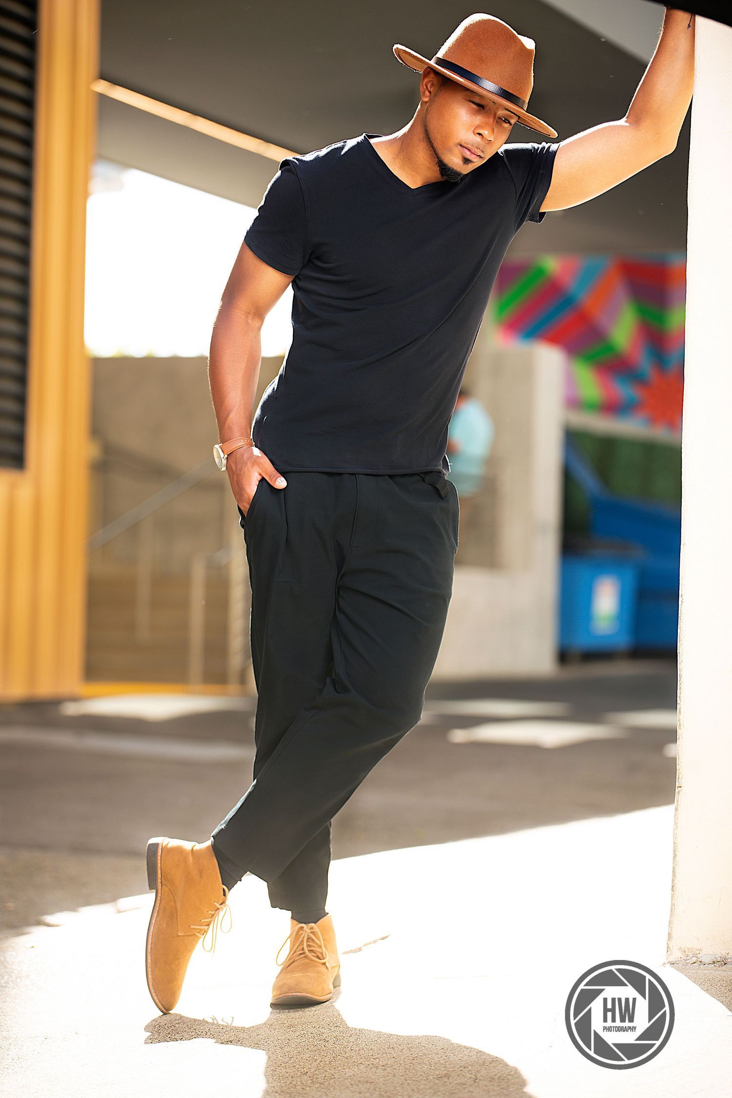 Stylish urban portrait of person in black t-shirt and brown hat leaning against colorful wall.