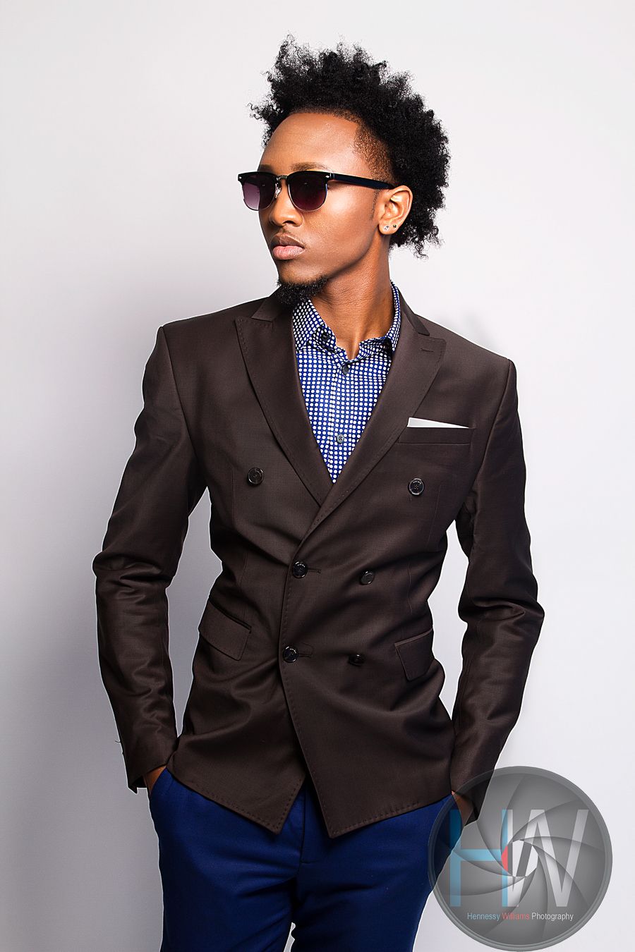 Fashion portrait featuring brown double-breasted blazer with checkered shirt and sunglasses against white backdrop.