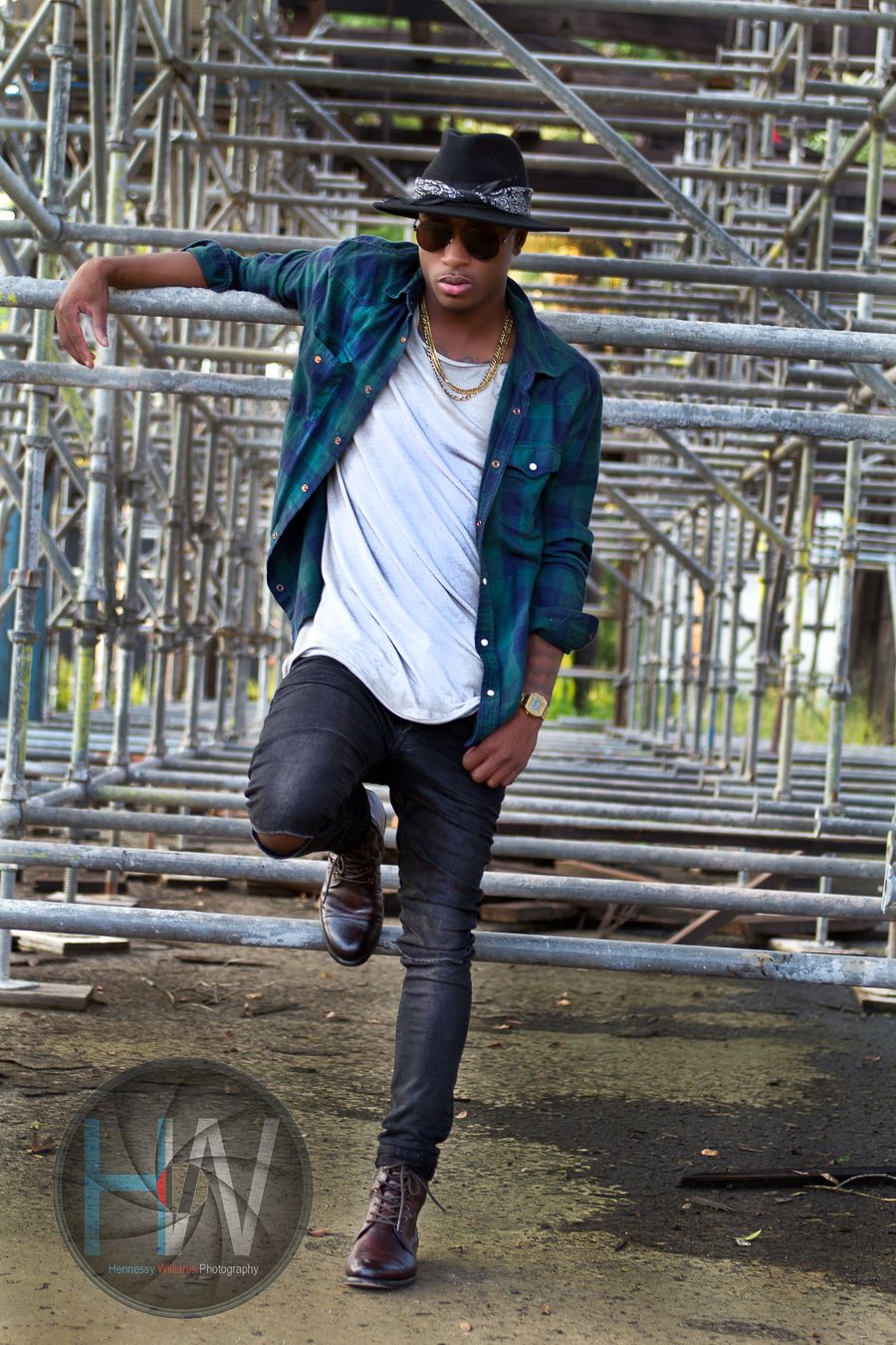 Person in casual street style with plaid jacket and sunglasses poses against industrial scaffolding background.