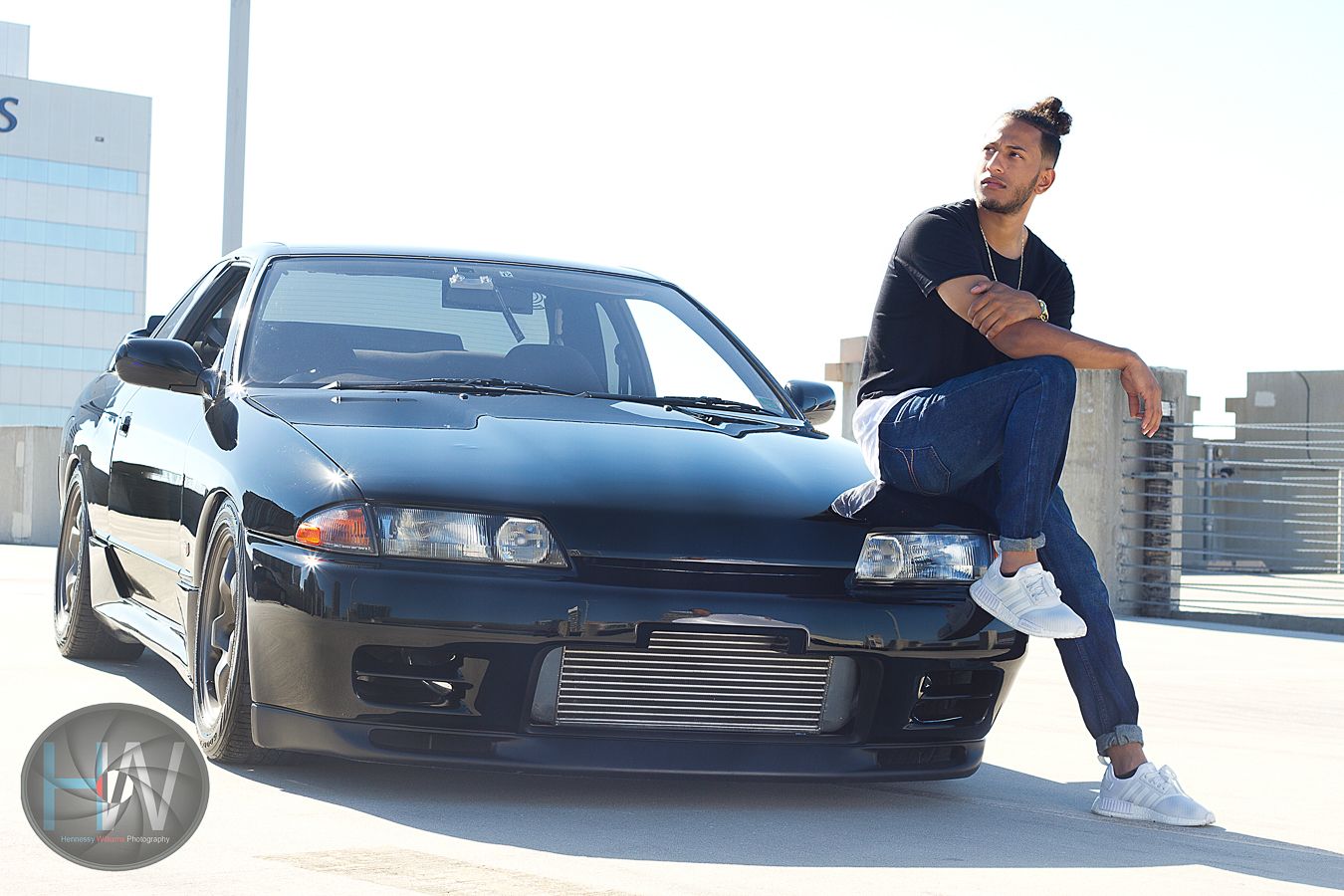 Lifestyle automotive shot with person leaning against black modified Nissan Skyline on urban rooftop.