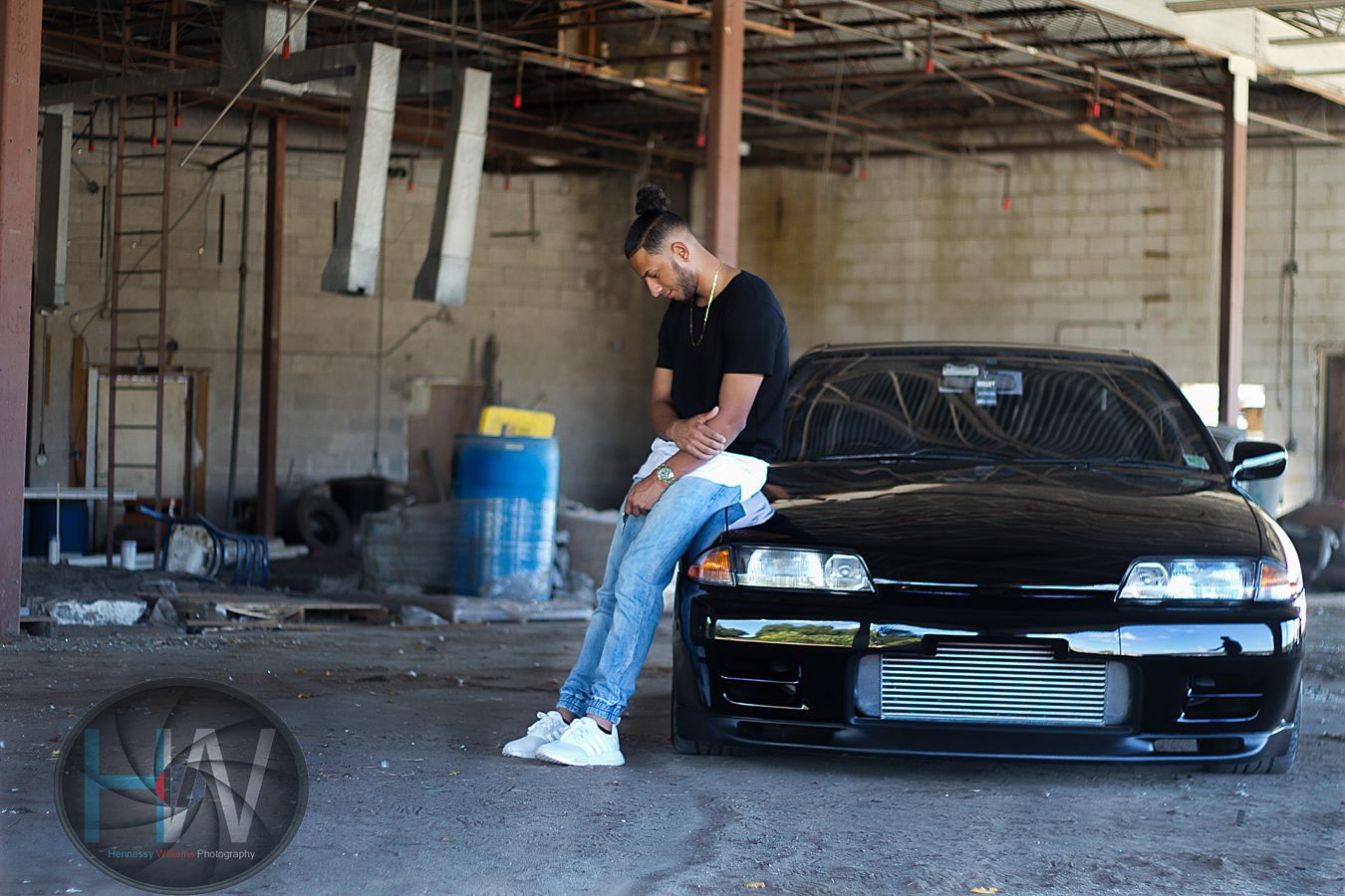 Person in black t-shirt sits on barrier next to modified black sports car in industrial setting.