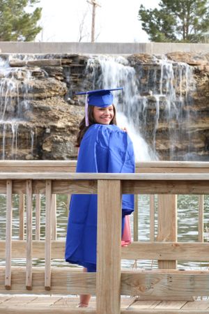 Graduate in blue cap and gown poses on wooden deck in front of cascading waterfall.