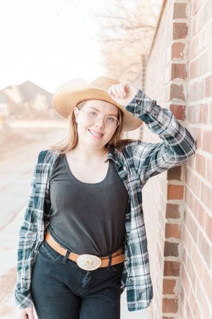 Western-inspired look with plaid shirt, tan hat and belt against brick wall in natural lighting.