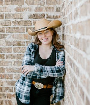 Western style portrait featuring plaid shirt, cowboy hat and belt against textured brick background.