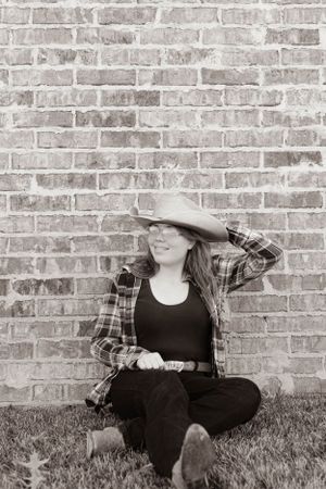 Black and white portrait of someone in western wear sitting on grass against brick wall.