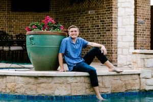 A casual outdoor portrait by a swimming pool with a large green planter and brick wall in the background.