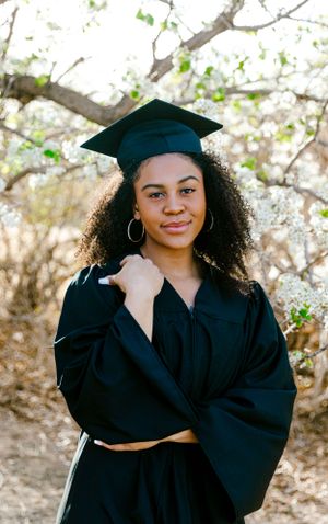 Graduate wearing academic regalia poses under flowering trees during graduation photo session.
