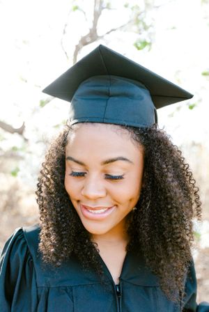 Graduate in cap and gown smiling with natural curly hair against blurred nature background.