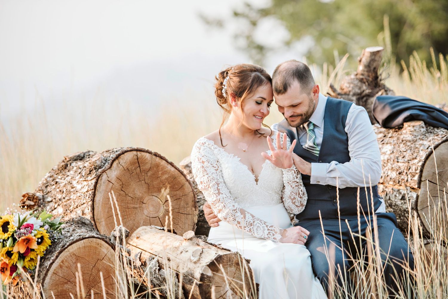 A bride and groom sit on logs while they admire the bride's ring