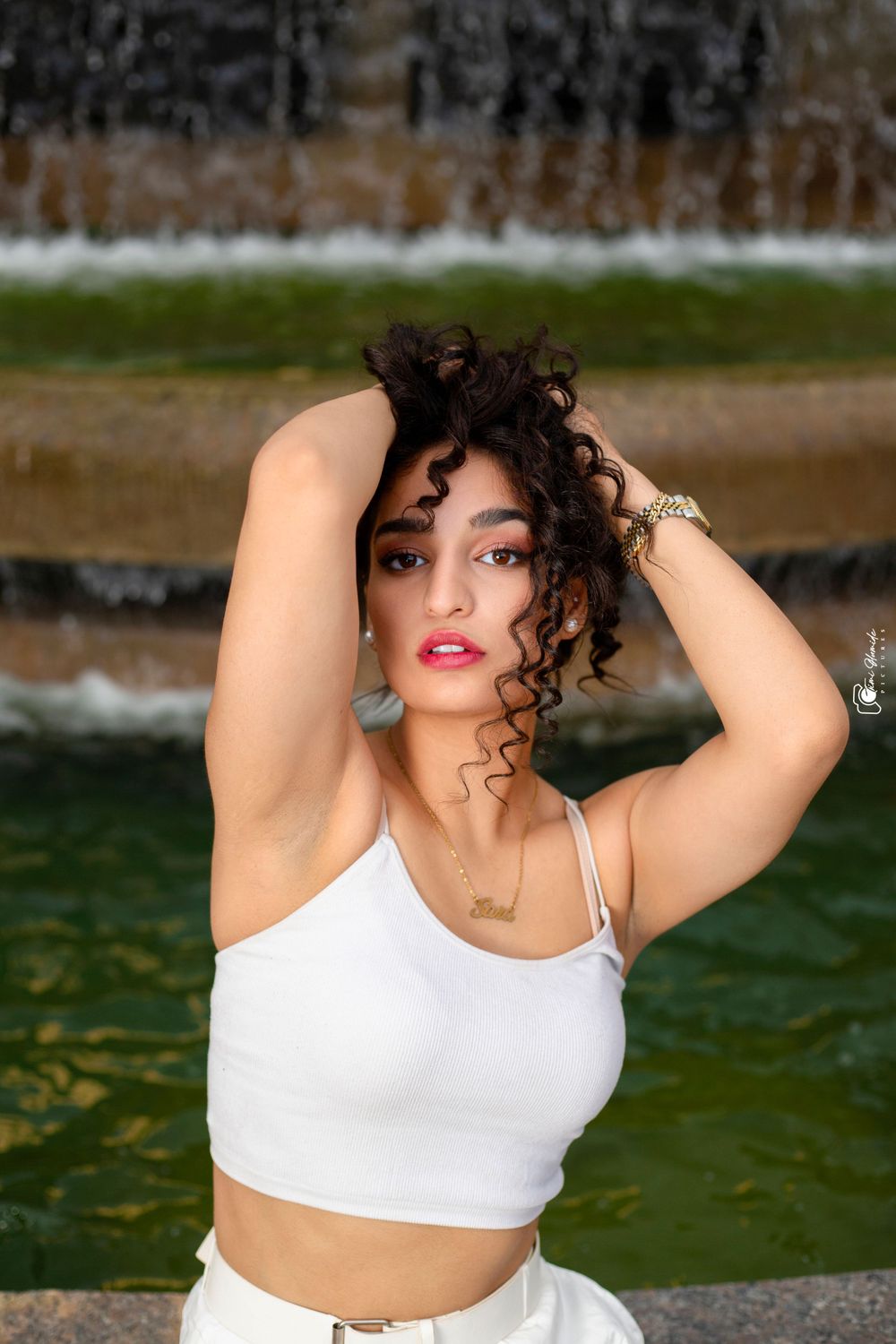Woman in white crop top poses outdoors near fountain with curly hair and red lipstick.