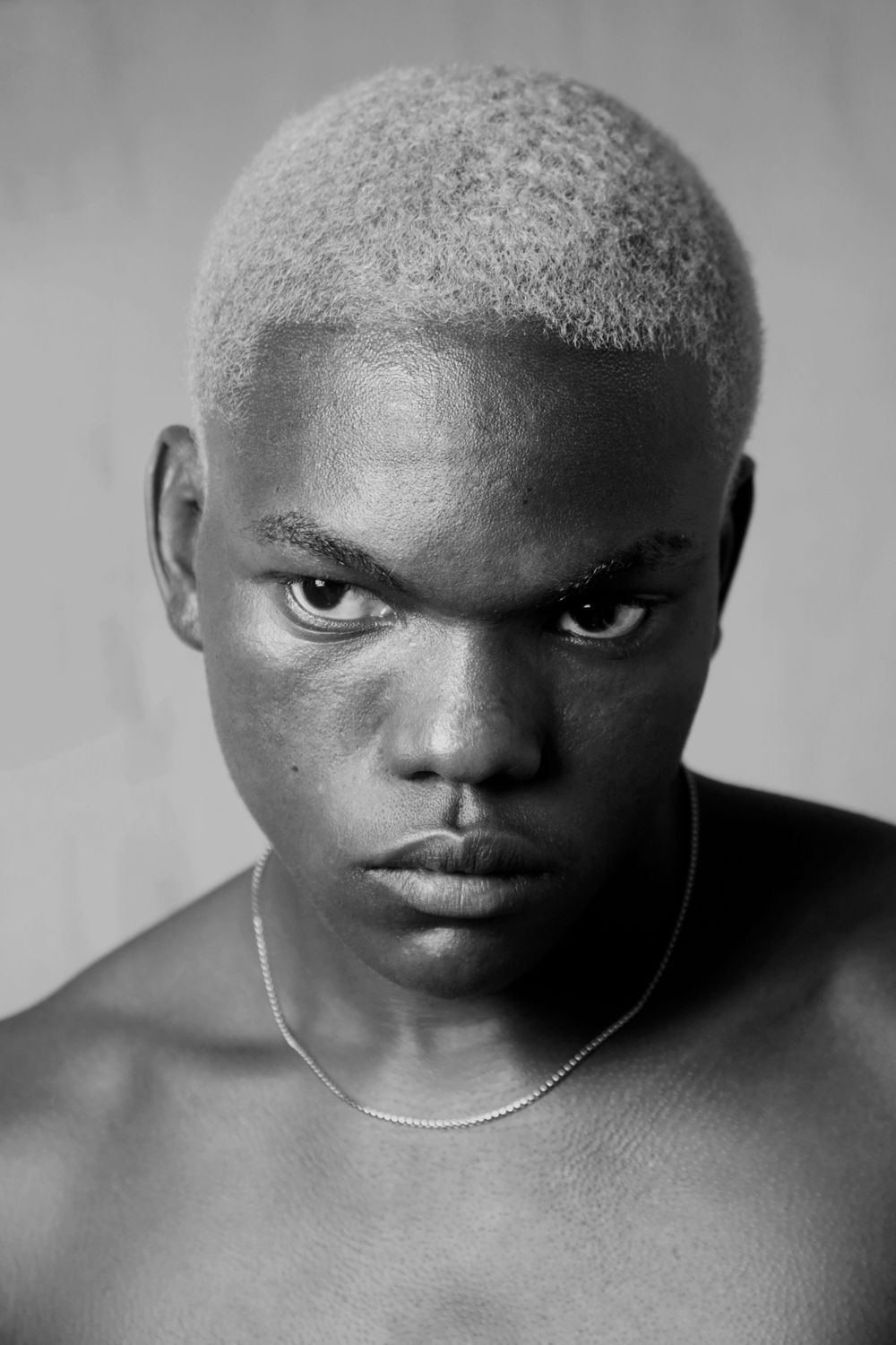 Black and white portrait with platinum blonde hair and silver necklace against neutral background.