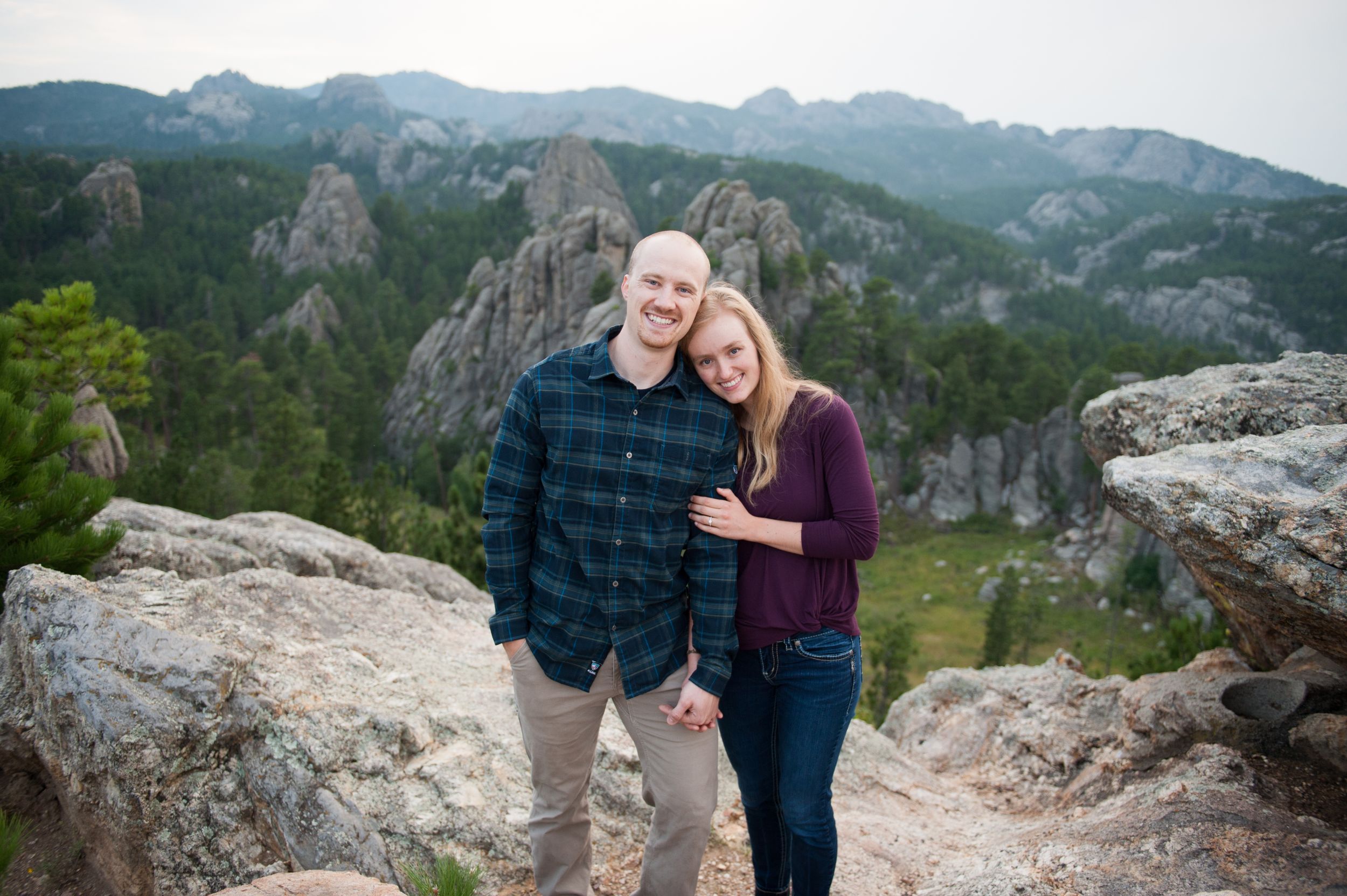 Couple poses on rocky mountain overlook with dramatic rock formations in background.