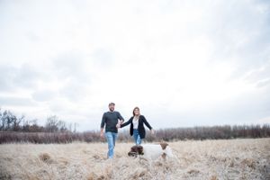 Couple walking together in a snowy winter field with bare trees in the background.