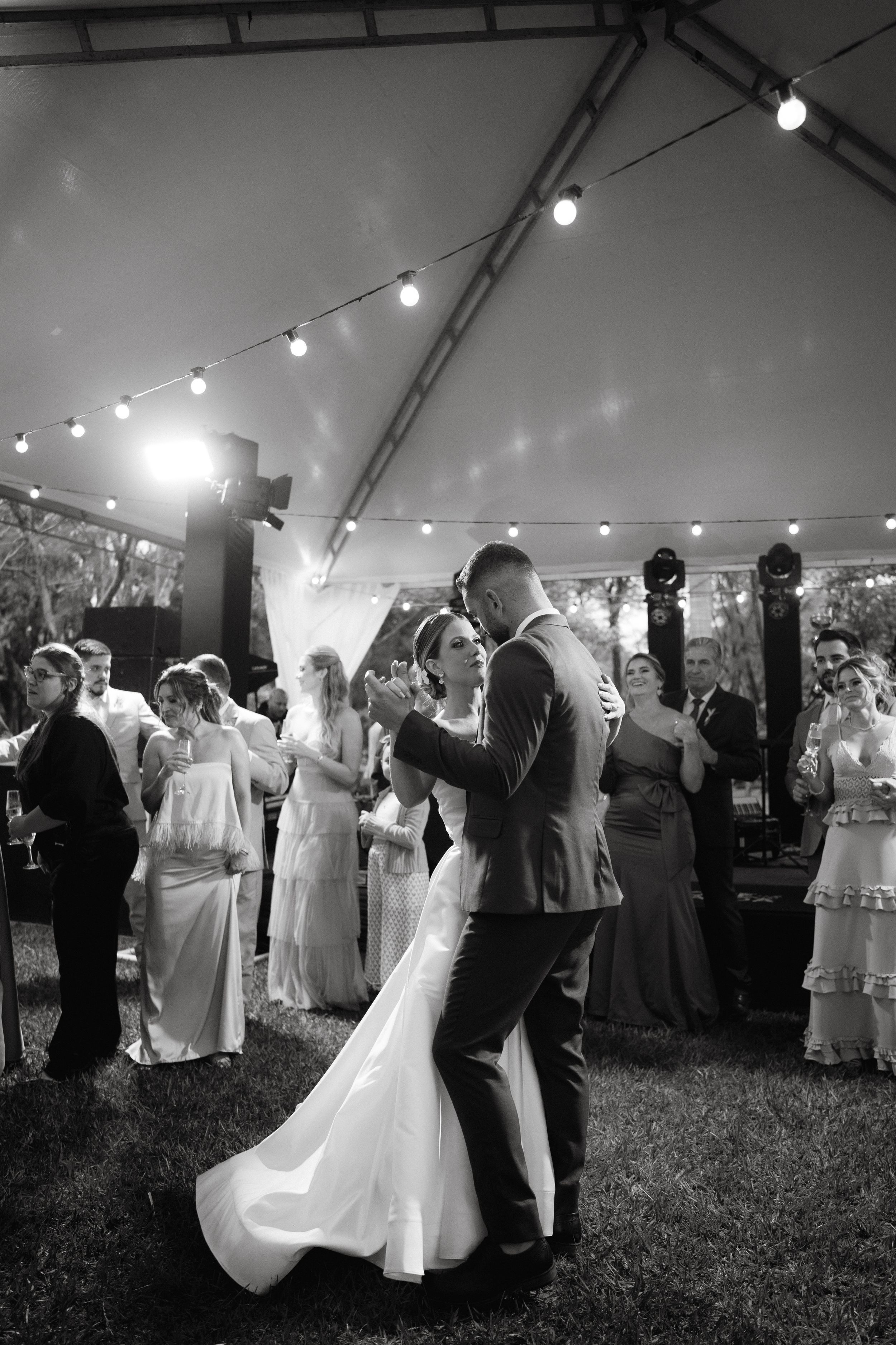 First dance under string lights at an outdoor wedding reception with guests watching.