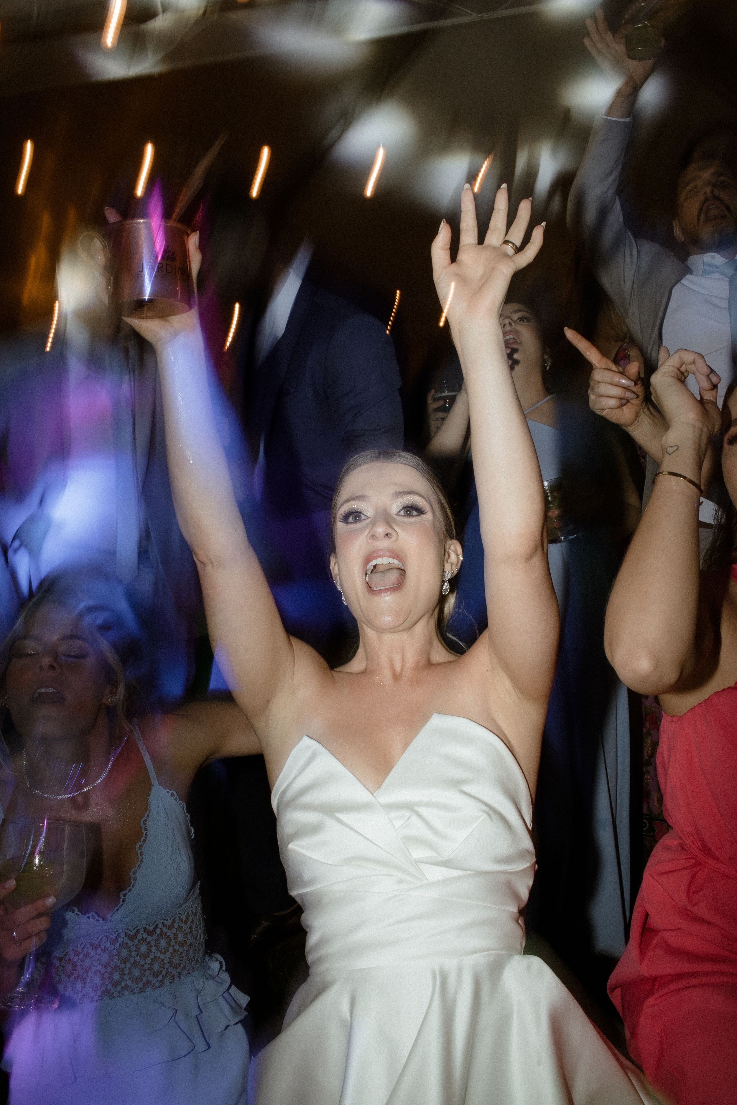 Bride in white dress dances joyfully with raised arms under colorful party lights at wedding reception.