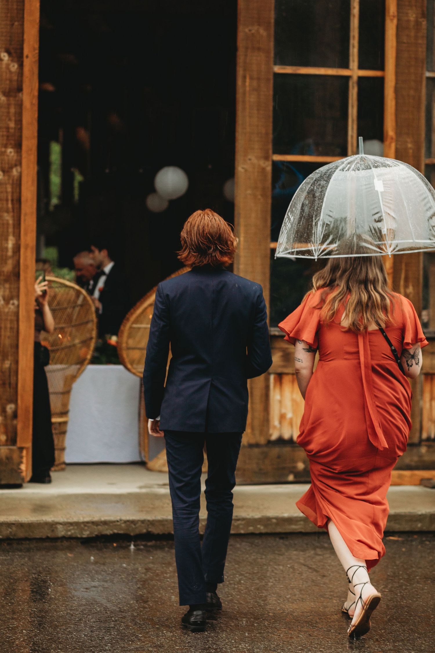 Couple walking in rain towards rustic venue entrance, with person in red dress holding clear umbrella.