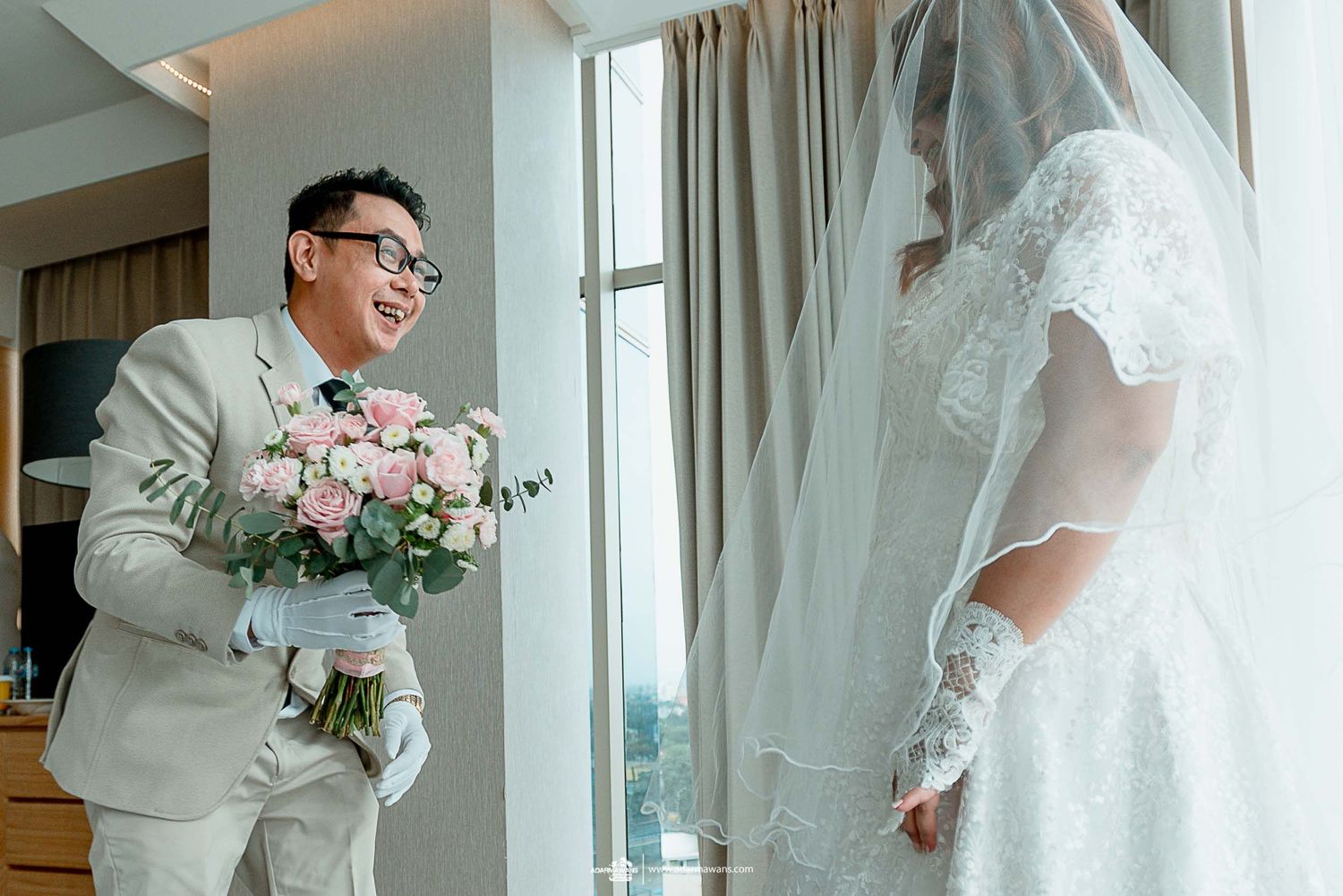 First look moment in a bright hotel room with pink floral bouquet.