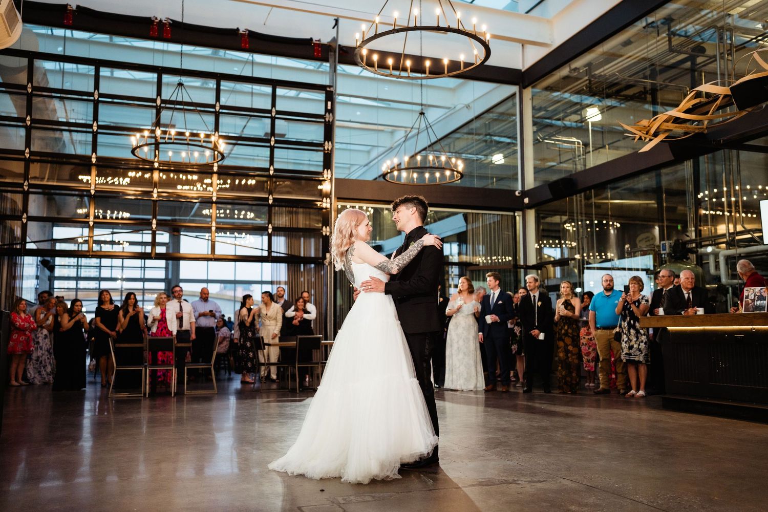 Wedding celebration with dancing couple in an industrial-style venue with high ceilings and chandeliers.