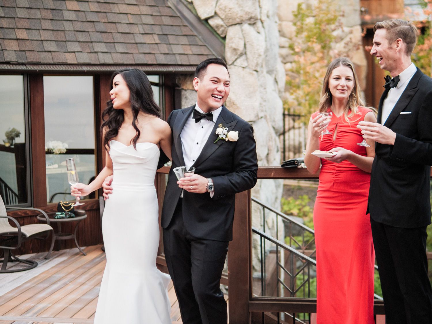 Wedding party in formal attire sharing drinks and laughs on an outdoor deck during a celebration.