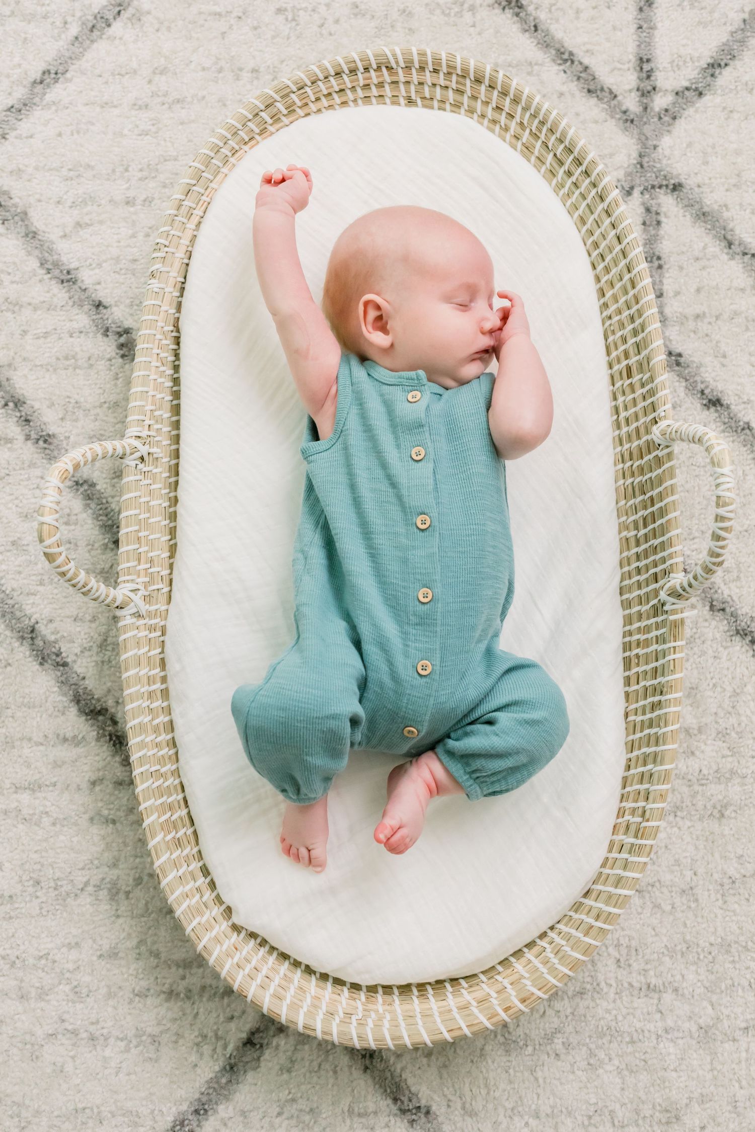 Infant in teal button-up romper stretching arms while resting in woven basket.