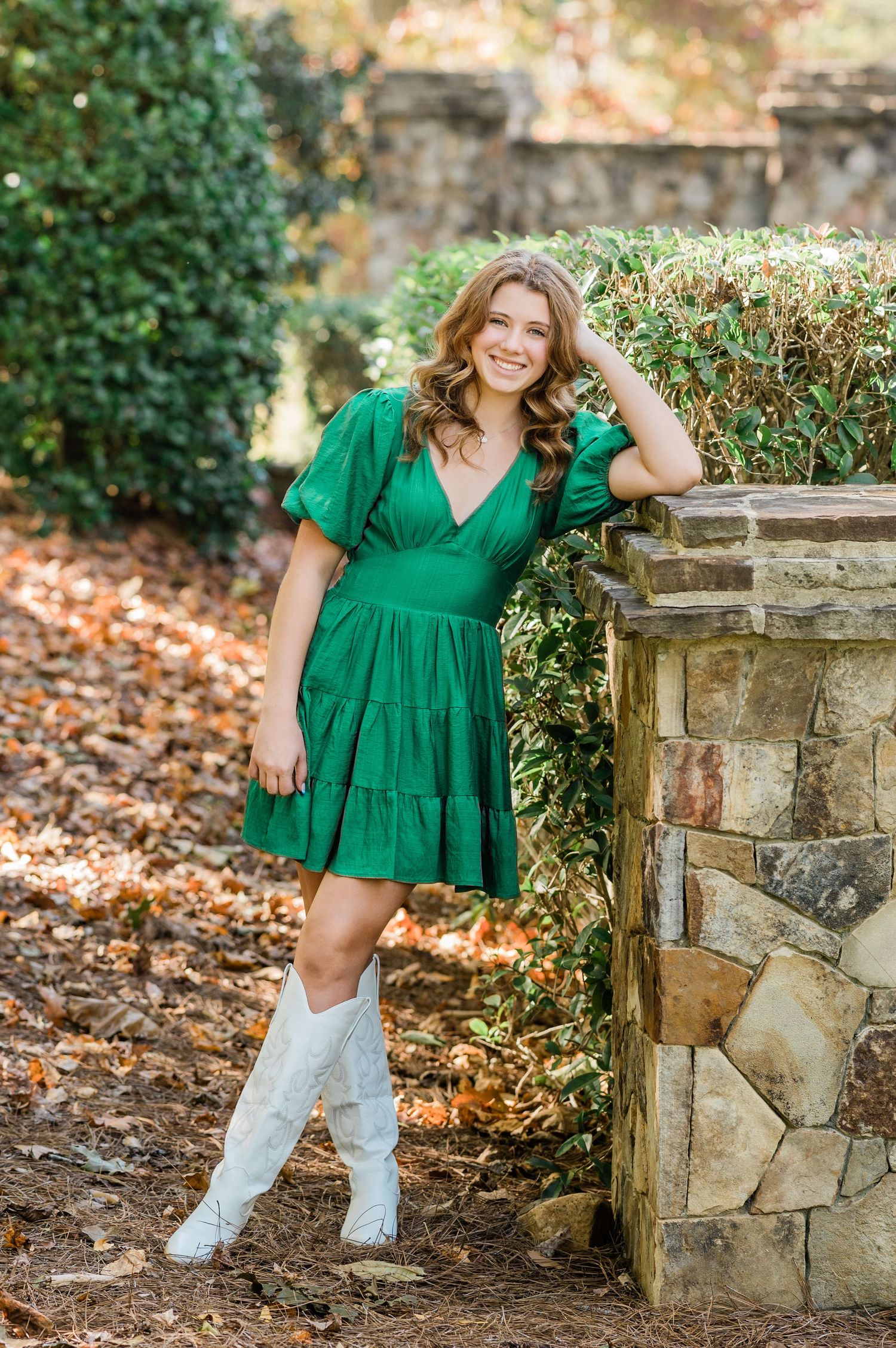 Outdoor photoshoot featuring green ruffle dress and white boots against stone wall backdrop with autumn leaves.