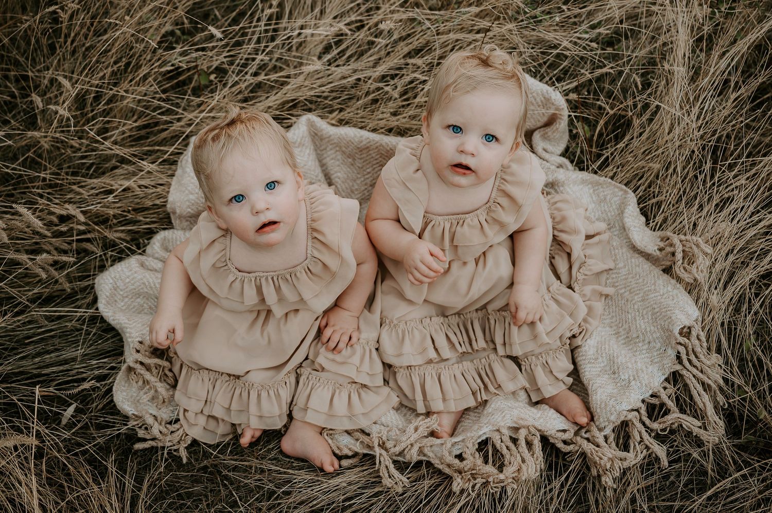 Two infants wearing matching ruffled dresses pose together on a blanket in tall grass.