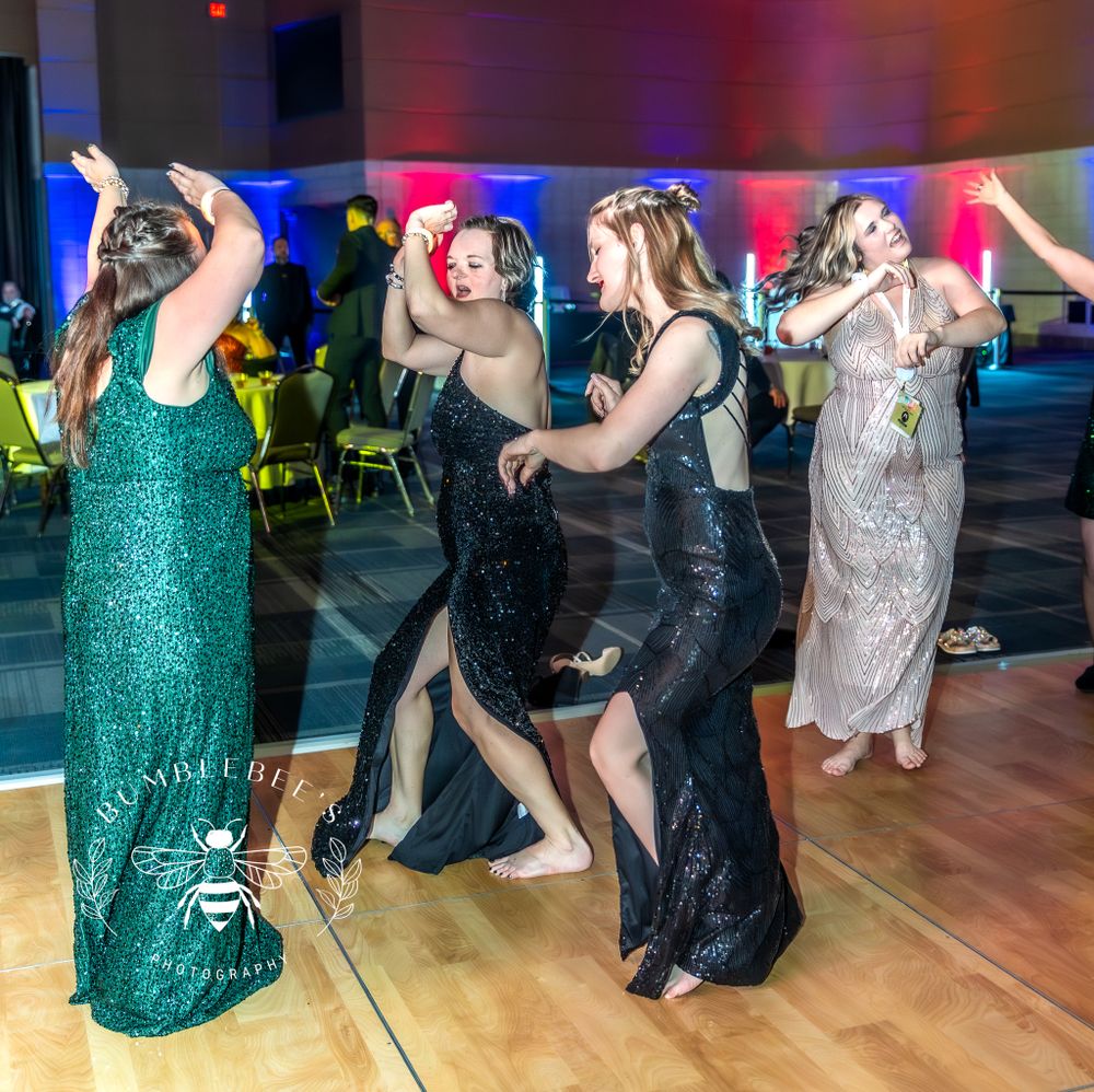 A group of people in formal dresses dance energetically at a party with colorful lighting and ambiance.