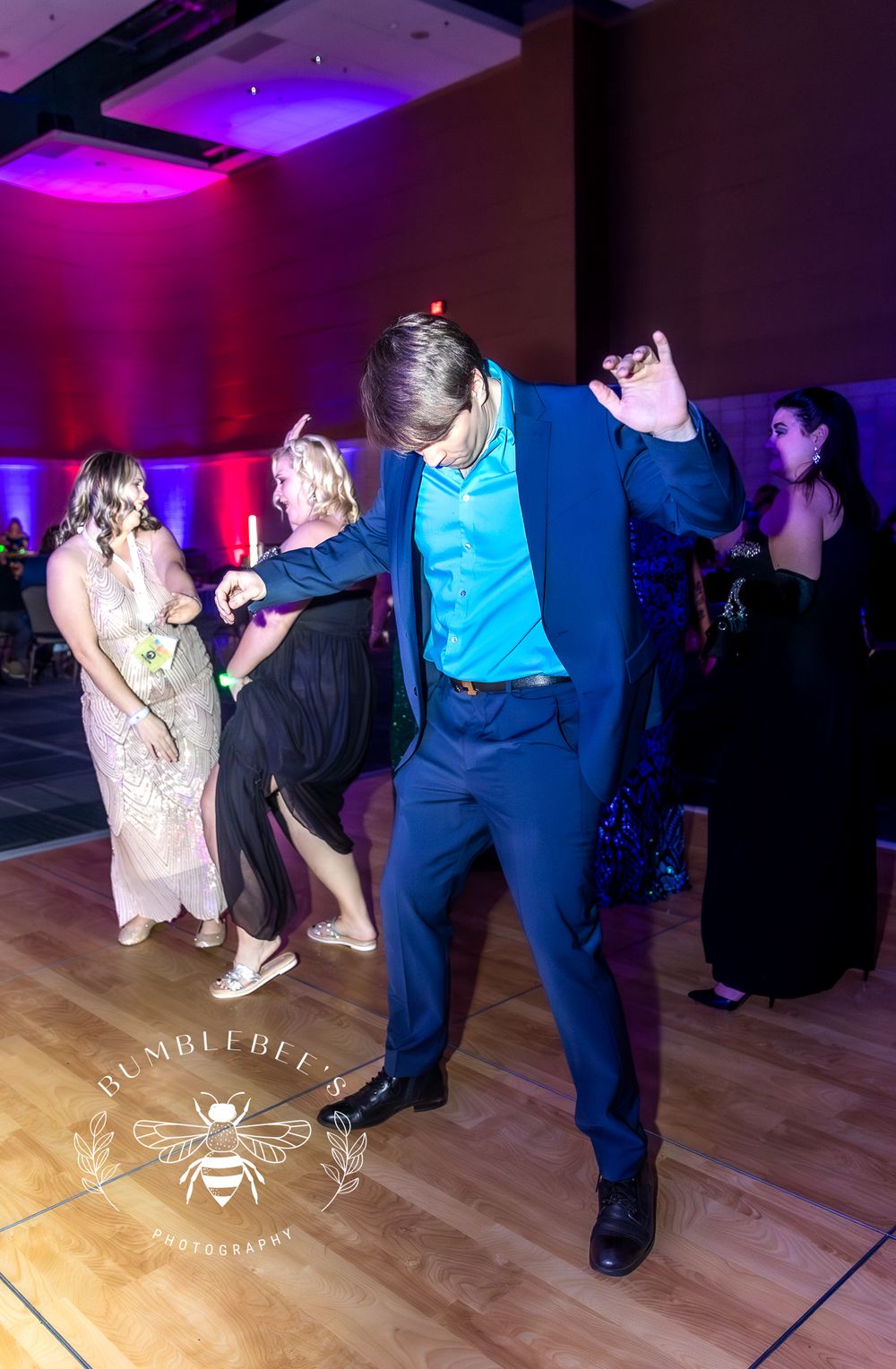 Person in blue suit enthusiastically dancing at formal event with colorful lighting.