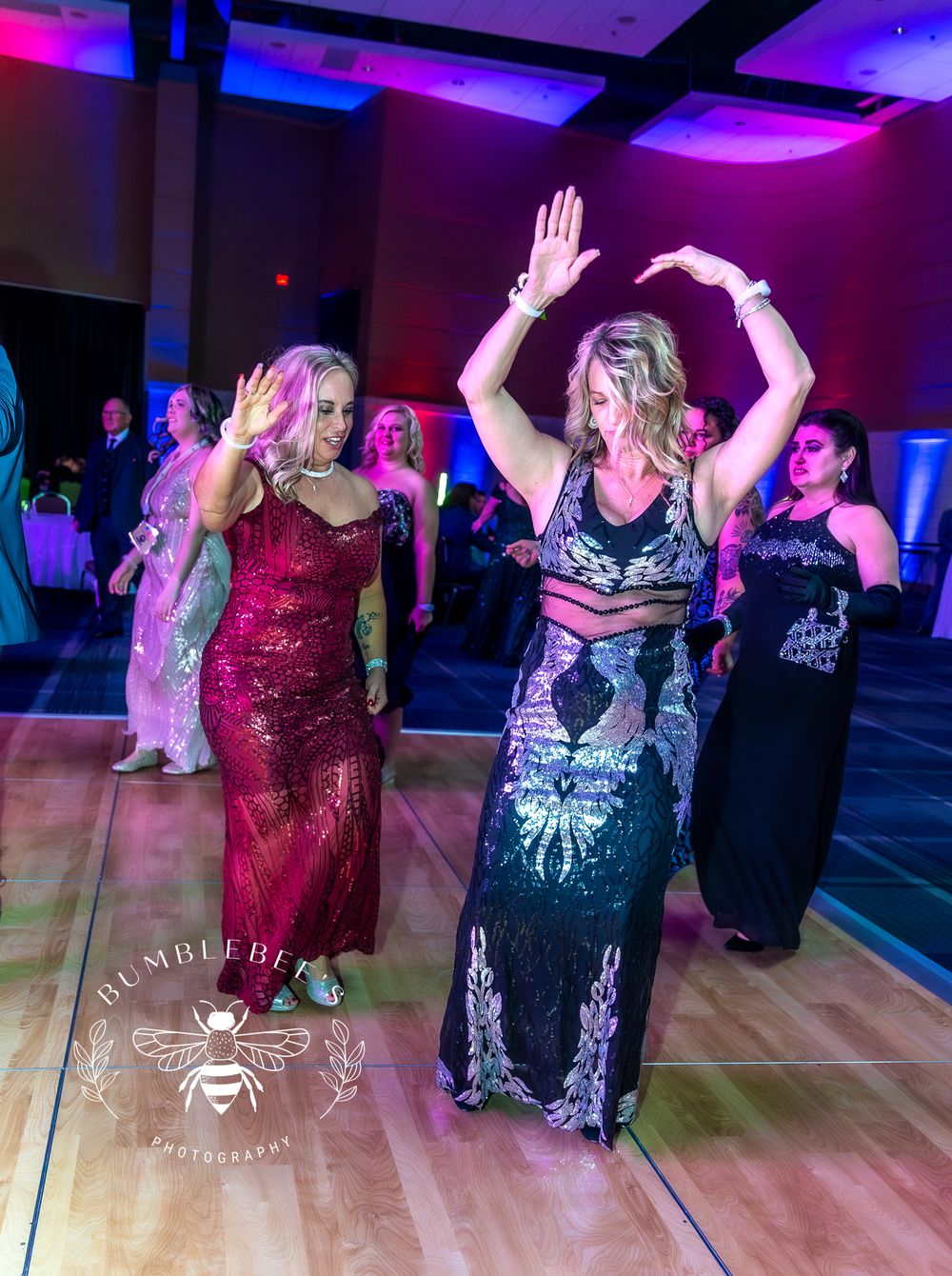People dancing and celebrating on wooden dance floor with purple and blue lighting effects.
