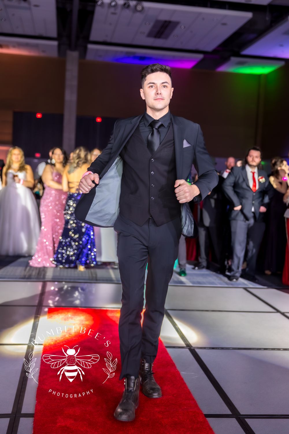 A formal fashion show participant walks confidently down a red carpet runway in an all-black suit.
