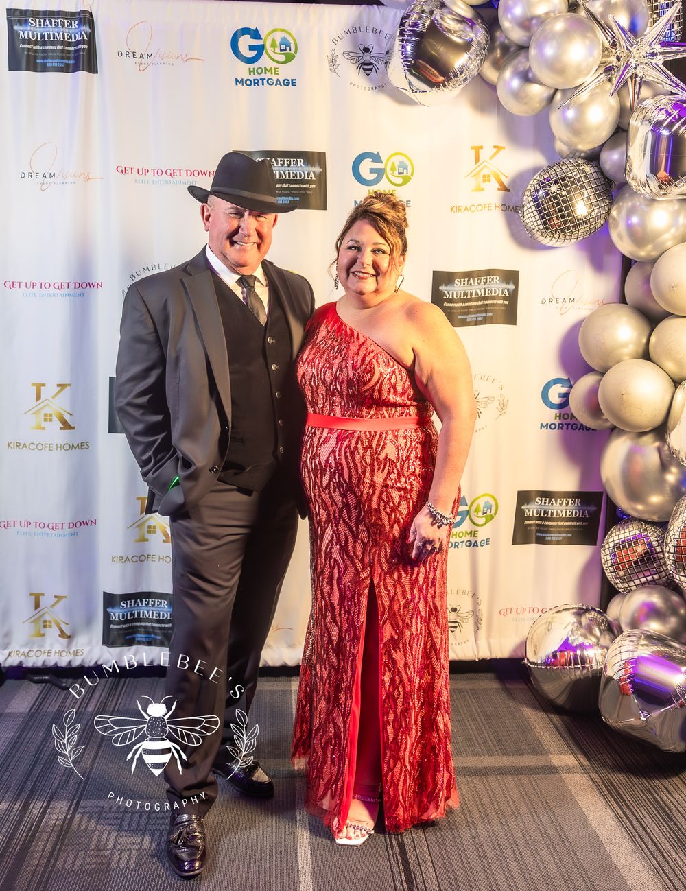 Guests pose at a formal event against a branded step-and-repeat backdrop with silver balloon decorations.