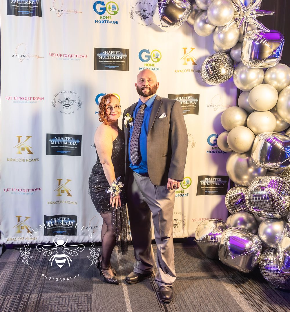 Formal event photo with party backdrop featuring silver and purple balloons and promotional banners.