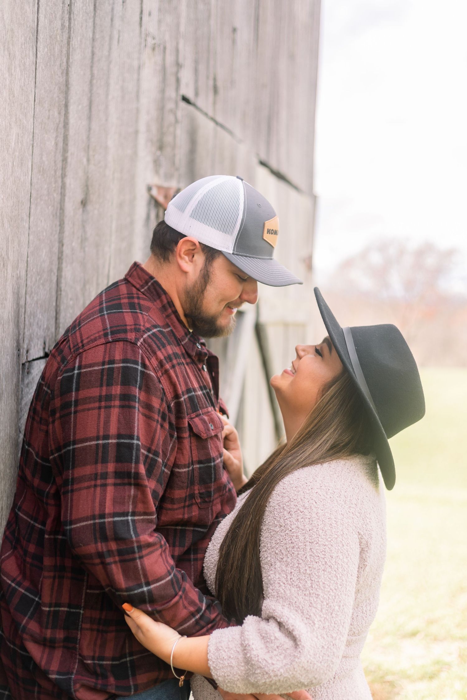 A couple in matching baseball caps and plaid share an intimate moment against a rustic barn wall.