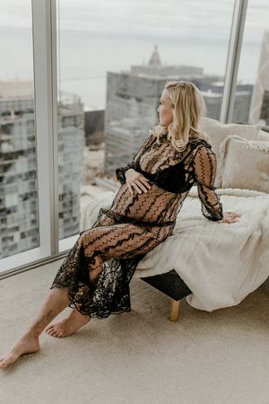 Relaxed pose in patterned dress by floor-to-ceiling windows with city views.