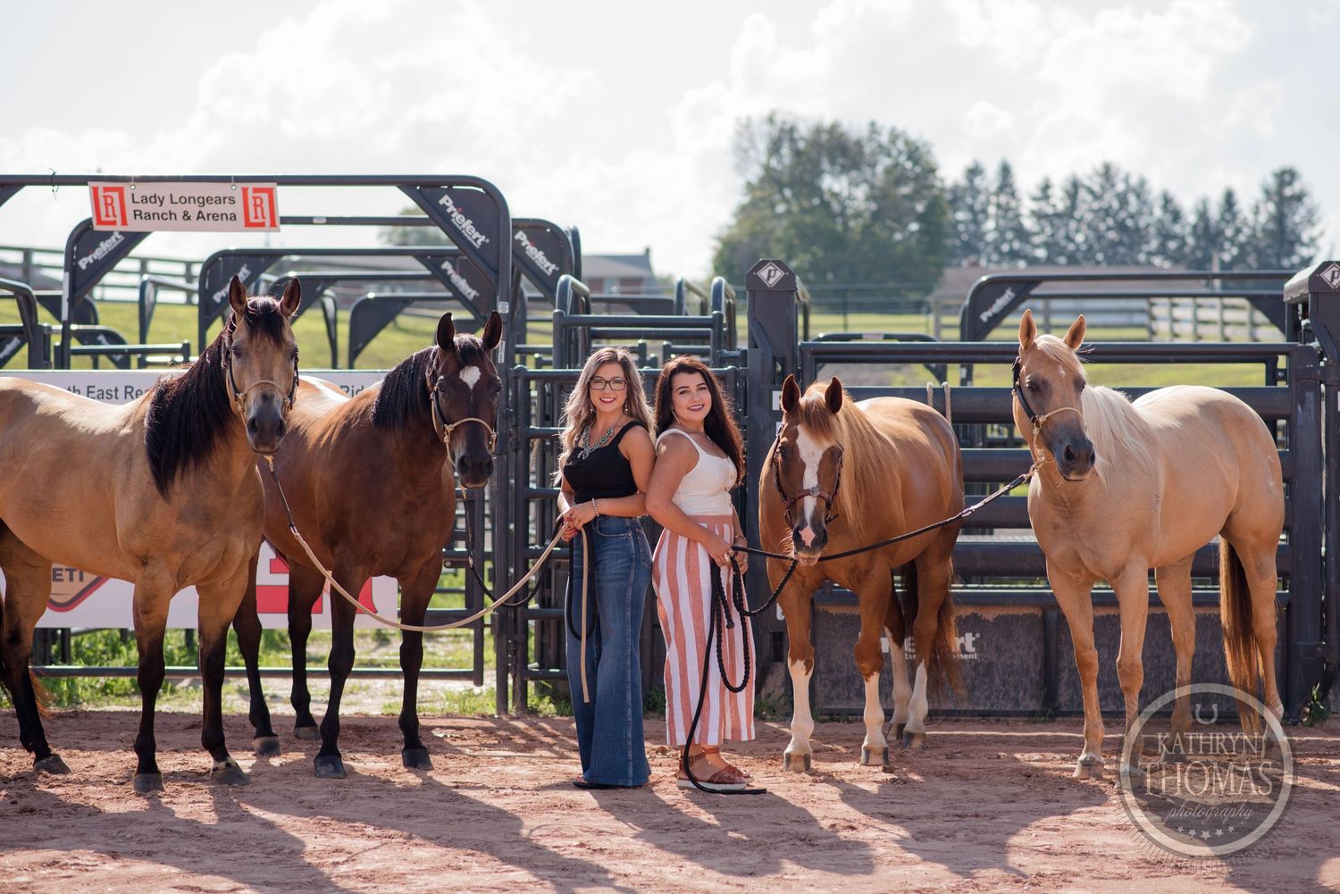 A group poses with several horses of different colors by metal gates at an outdoor equestrian facility.