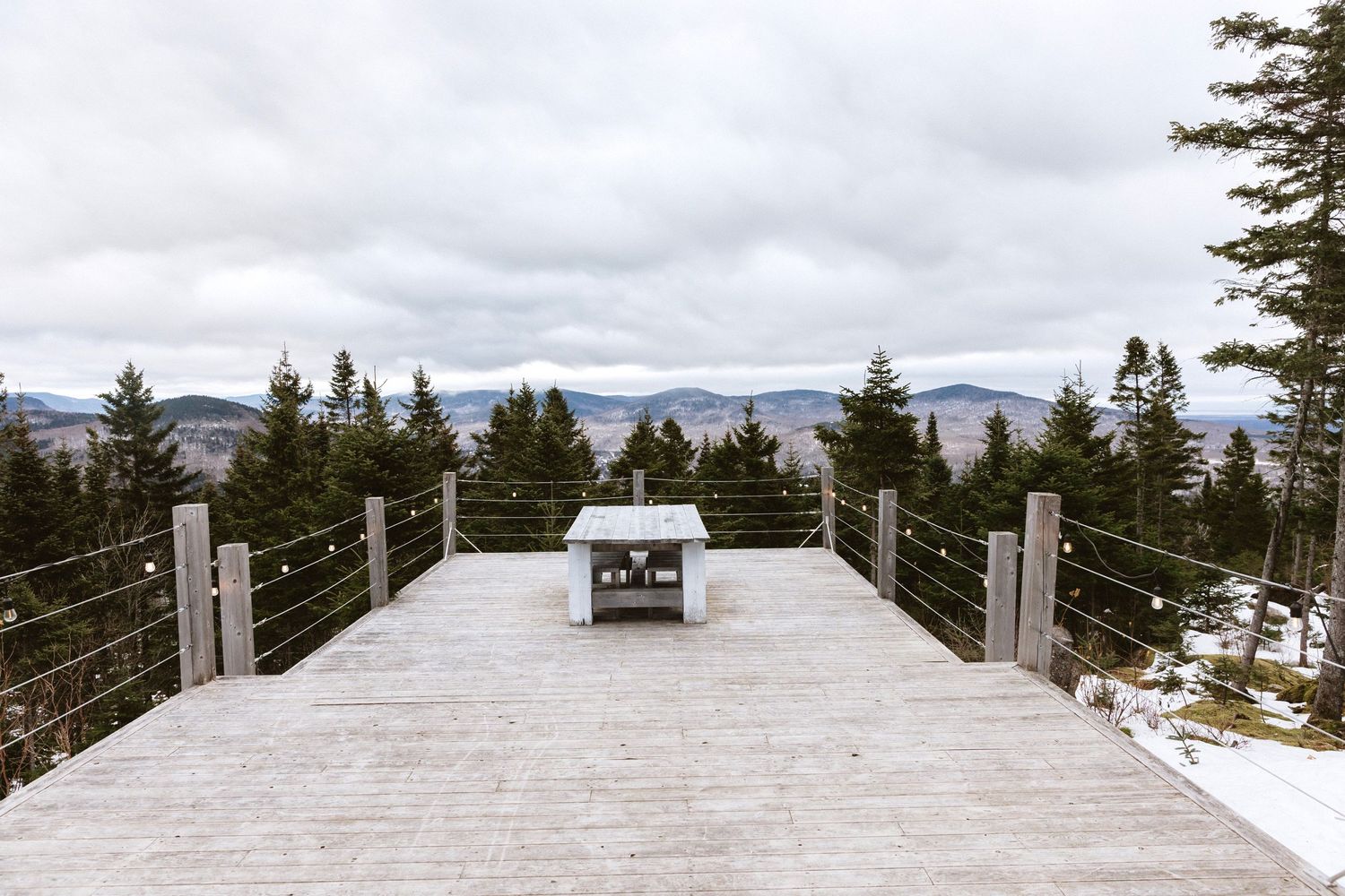 Snow-covered wooden deck overlooking mountain vistas with evergreen trees in the foreground.