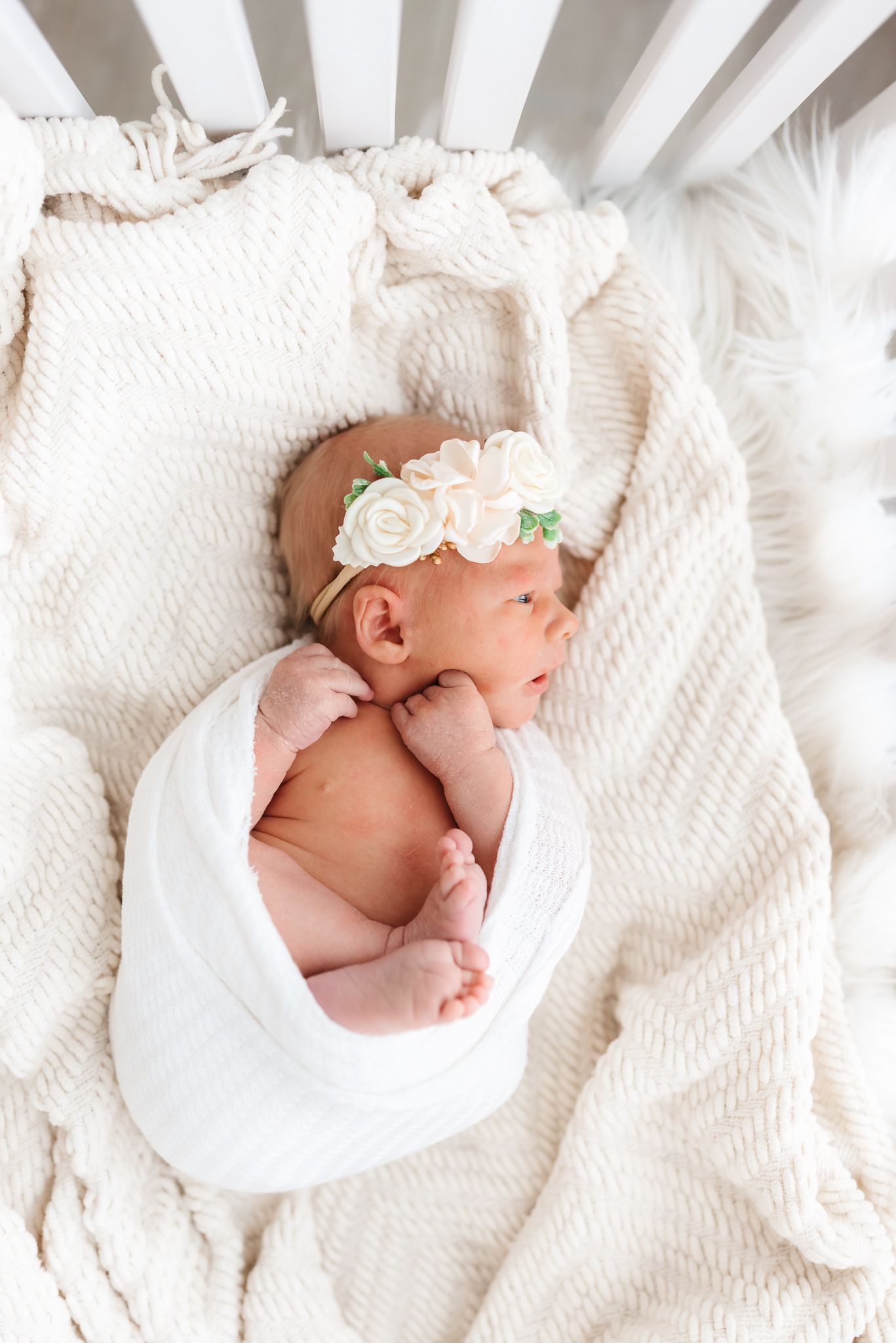 Newborn baby wrapped in white swaddle lying on textured cream blanket with delicate floral headband.