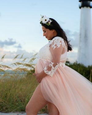 Pregnant woman in pink tulle maternity gown and floral crown posing near beach lighthouse.
