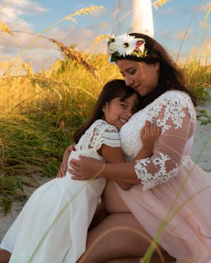 Mother in floral crown and lace dress embracing young daughter on beach at sunset.