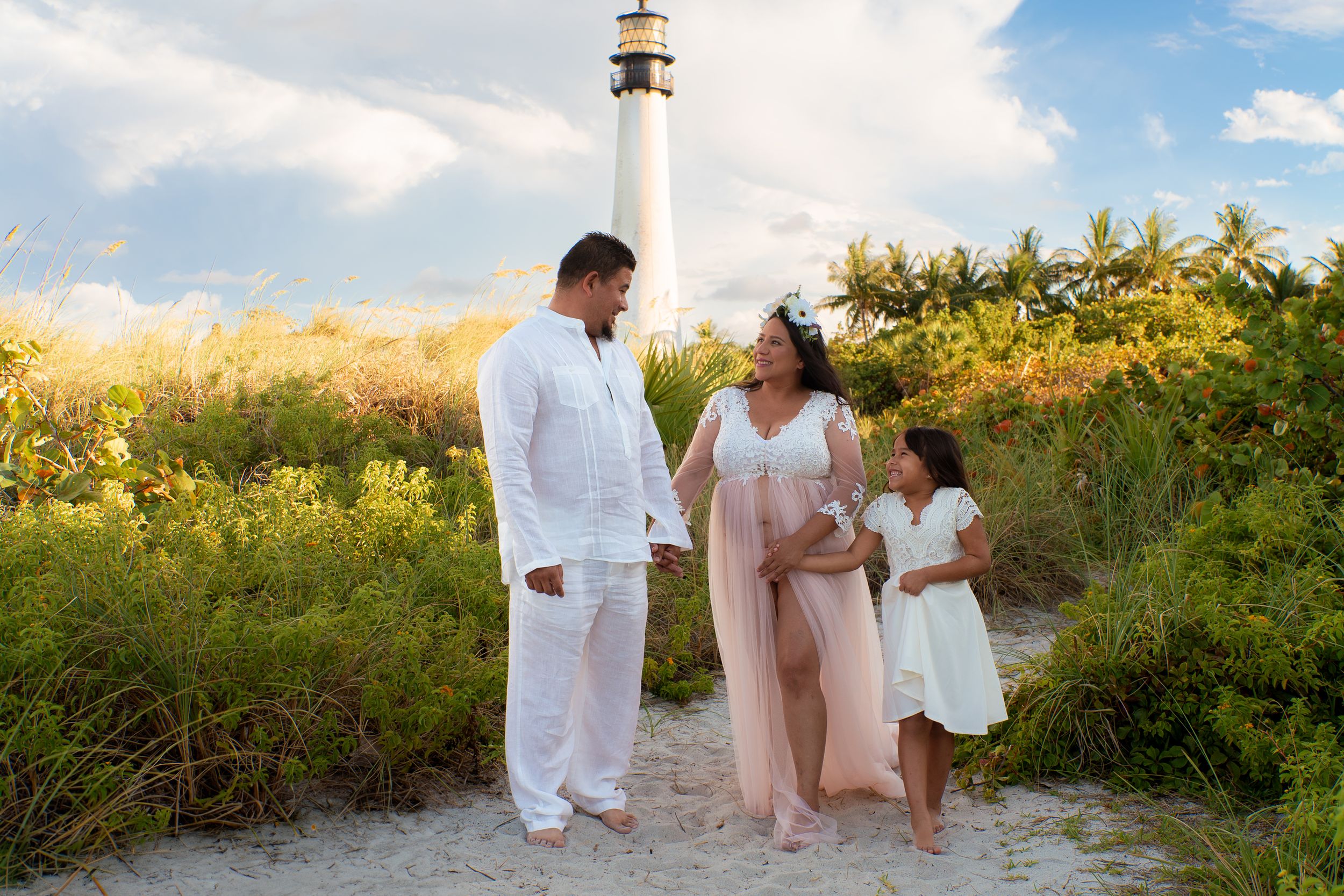 Pregnant woman in flowing pink gown walks with partner and daughter near Cape Florida lighthouse at golden hour.