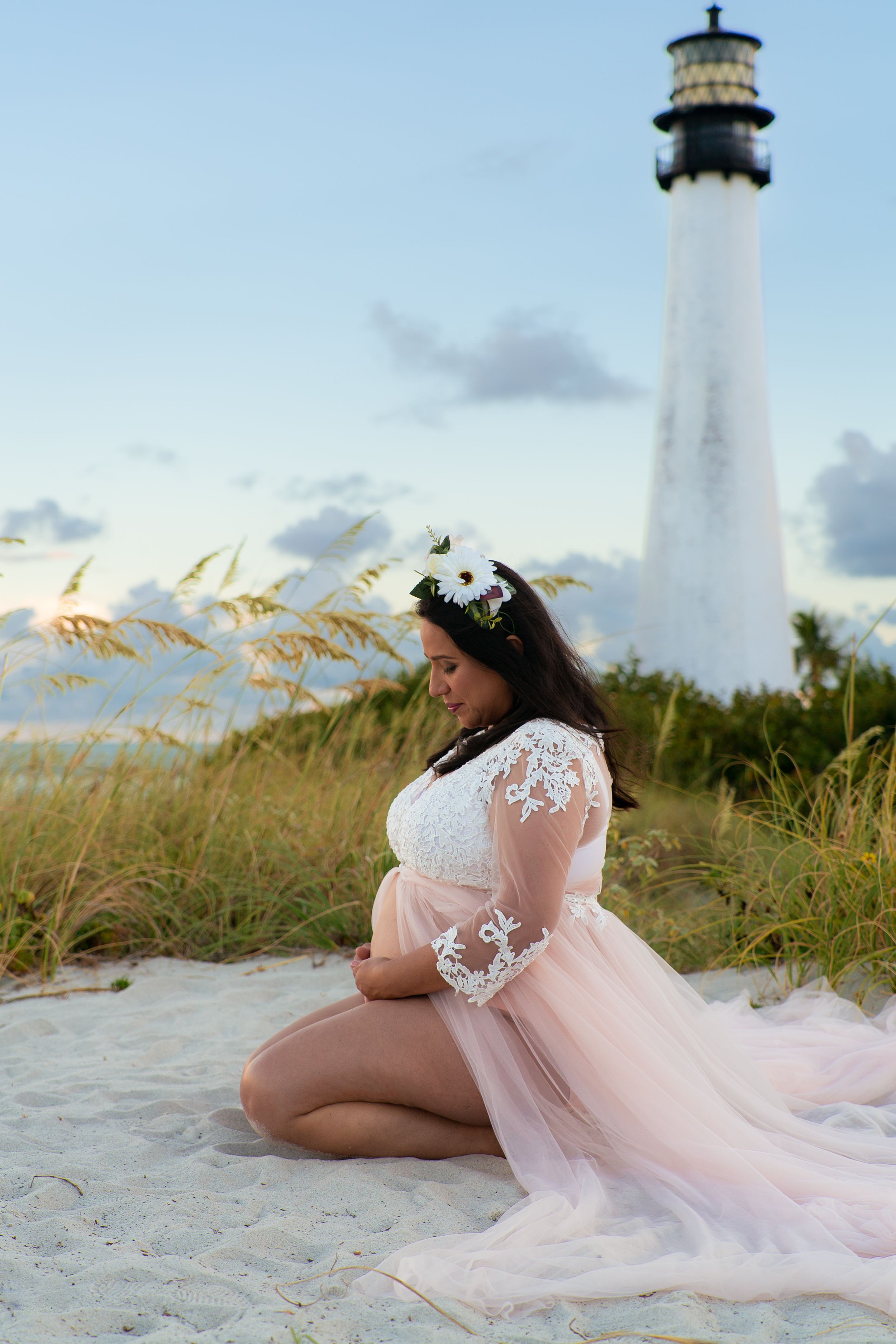 Pregnant woman in lace and pink tulle gown kneeling on sand before coastal lighthouse.